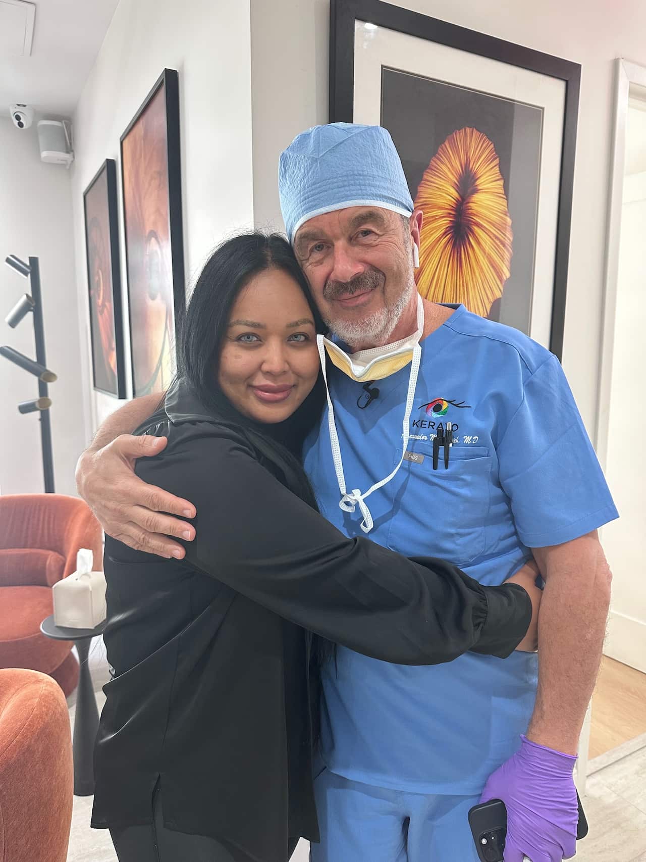 A woman with green eyes and a black jumper is hugging a man dressed in blue scrubs and a beard.