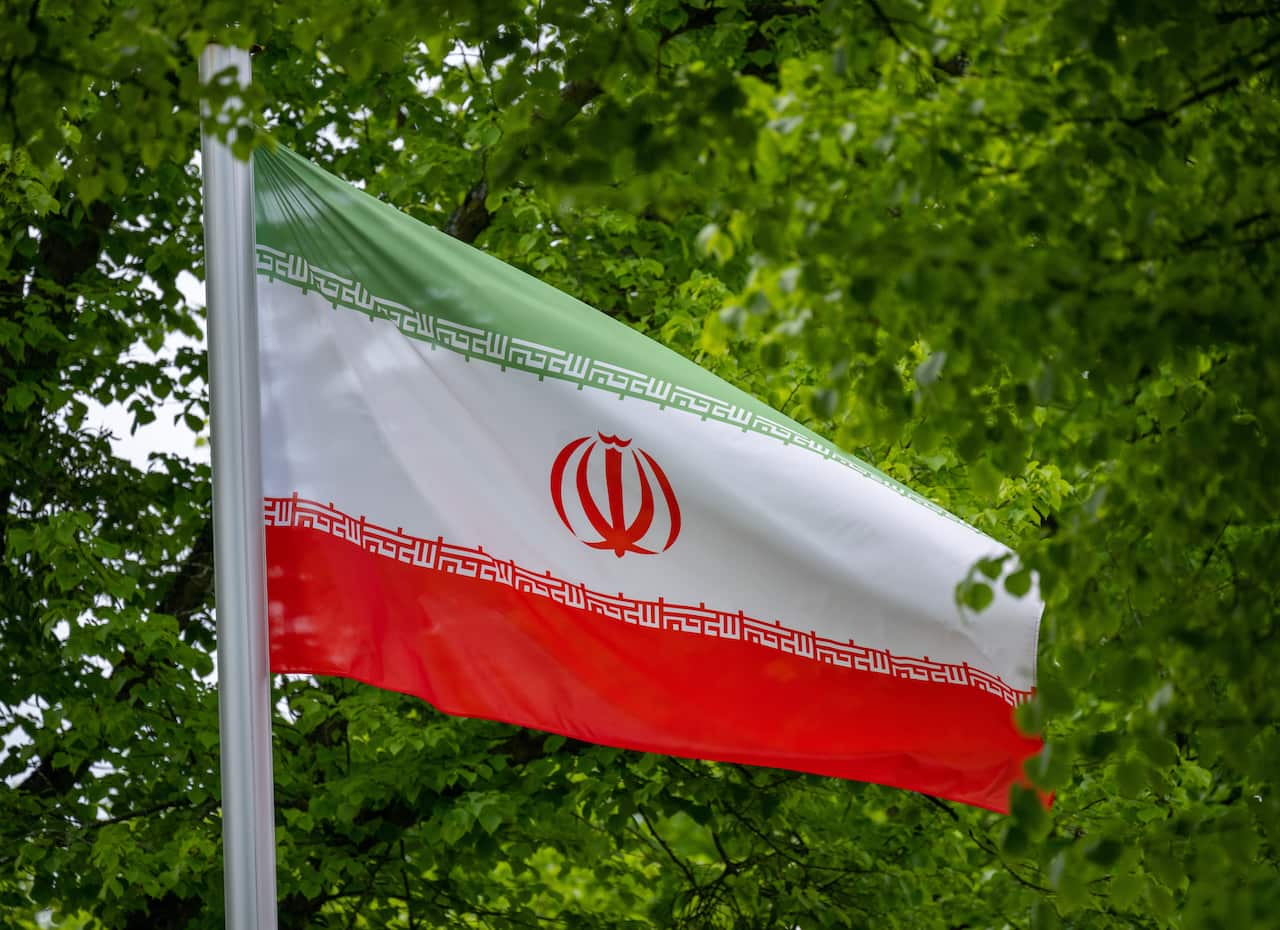 The national flag of Iran, featuring green, white, and red horizontal stripes with the central red emblem and stylized Kufic script, waves against a backdrop of lush green leaves.