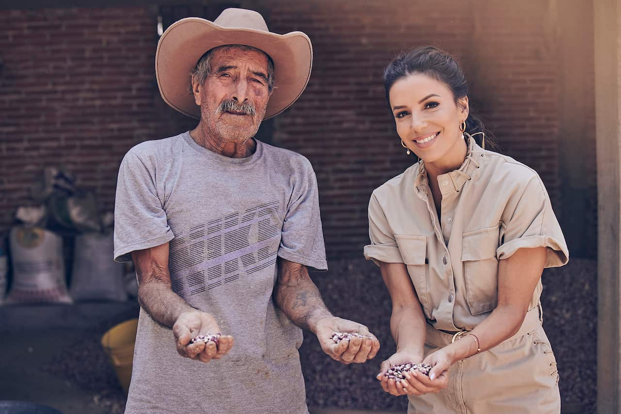 Un hombre con bigote gris y un sombrero de ala ancha estilo occidental y una mujer sonriente con un traje marrón están de pie con las manos llenas de maíz. 