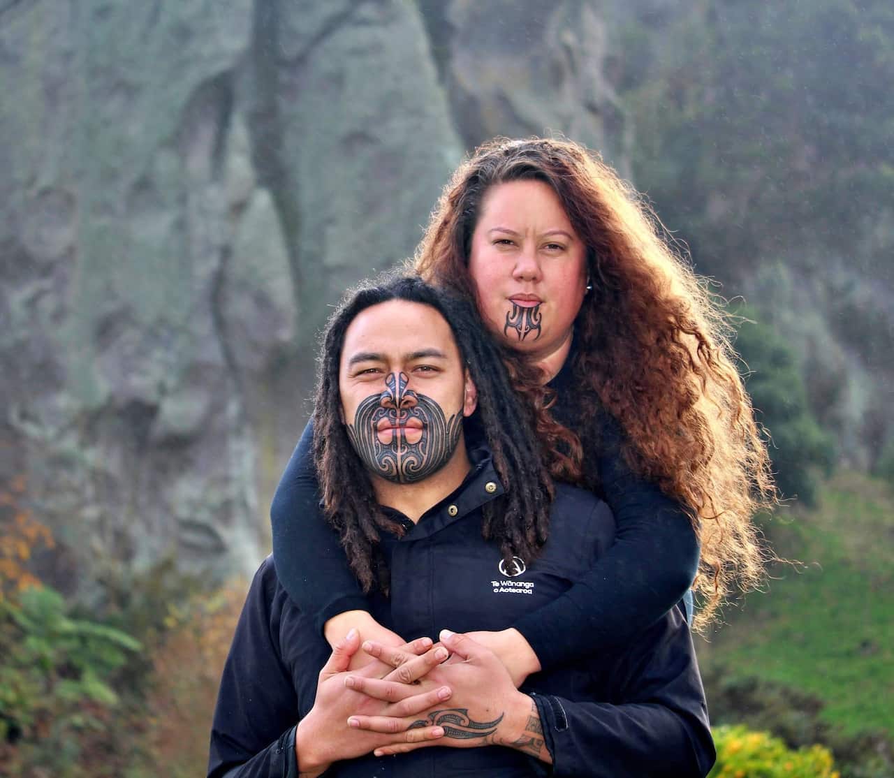 A man and a woman facing the camera. The woman is embracing the man from behind. The man has a traditional tattoo on the lower half of his face and the woman has a traditional tattoo on her chin. Mountains and greenery are in the backdrop.