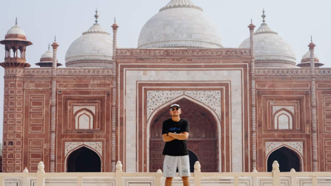 a young man in shorts, tshirt, sunglasses and a cap stands crossed-arm smiling in front of the Taj Mahal
