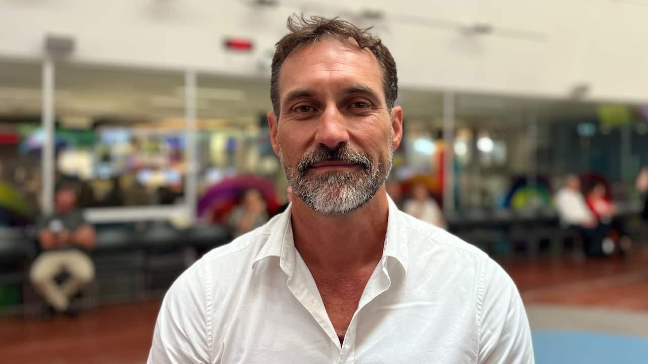 A close-up of a man with salt and pepper hair and beard in a white collared shirt, with an office in the background