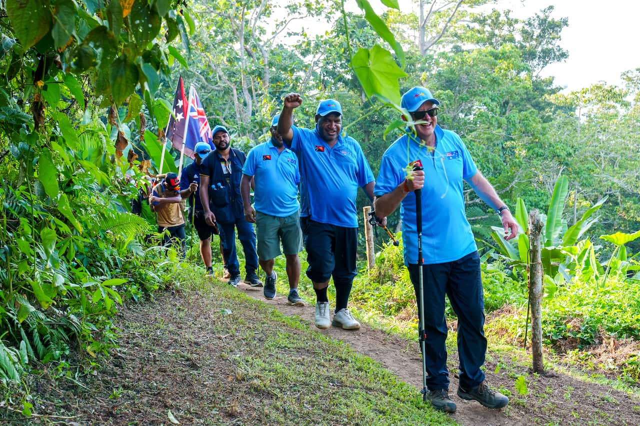 A row of men in light blue shirts. Some are wearing shorts and others long pants. They are walking in single file along a dirt track