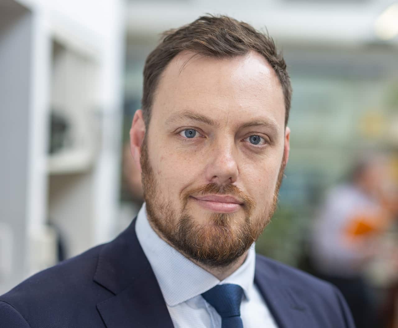 A head-and-shoulders portrait of a fair-skinned man with short brown hair and a beard, wearing a navy suit jacket, light blue shirt, and dark tie against a blurred office background.