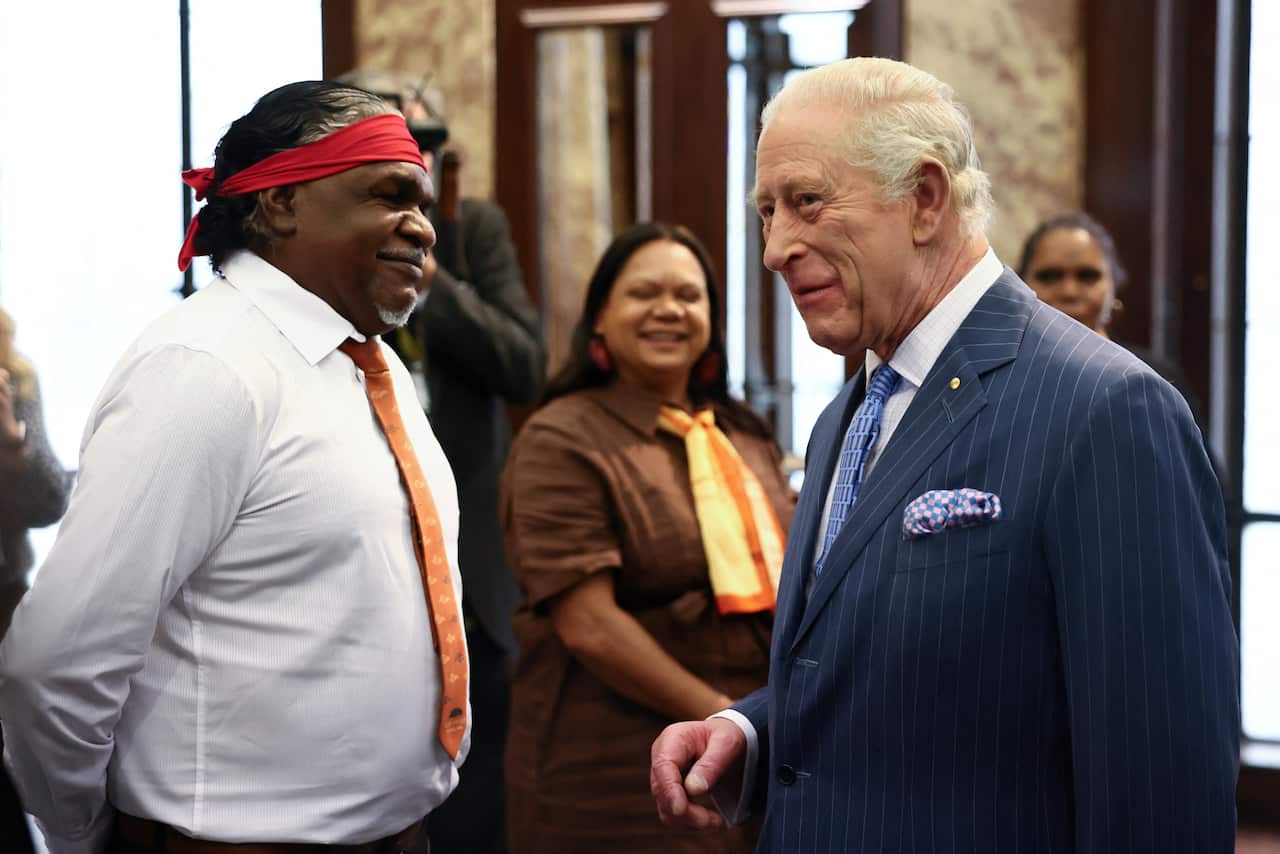 An Aboriginal man wearing a red head scarf, white shirt and tie greeted by King Charles, who is wearing a navy pin-stripe suit