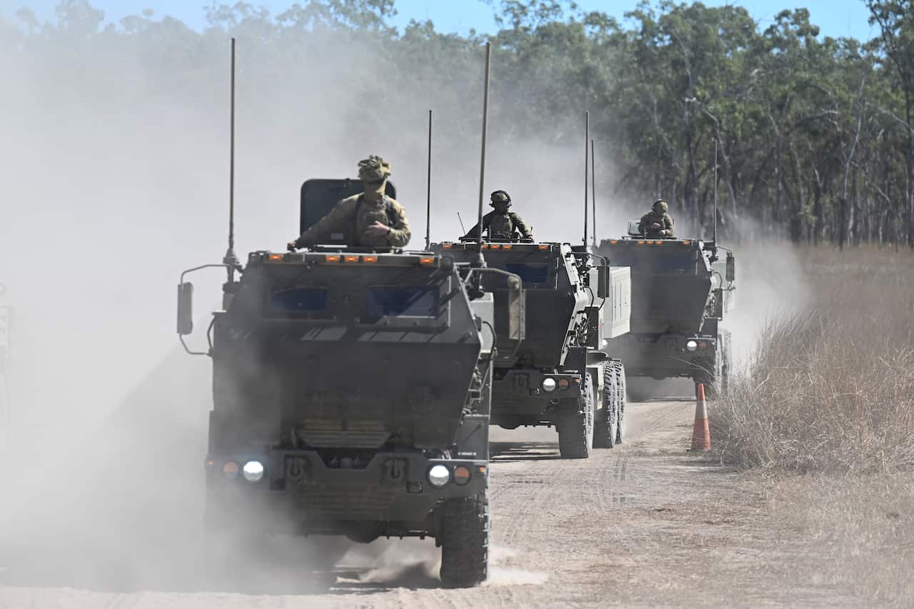 Three military trucks driving along a dirt road 