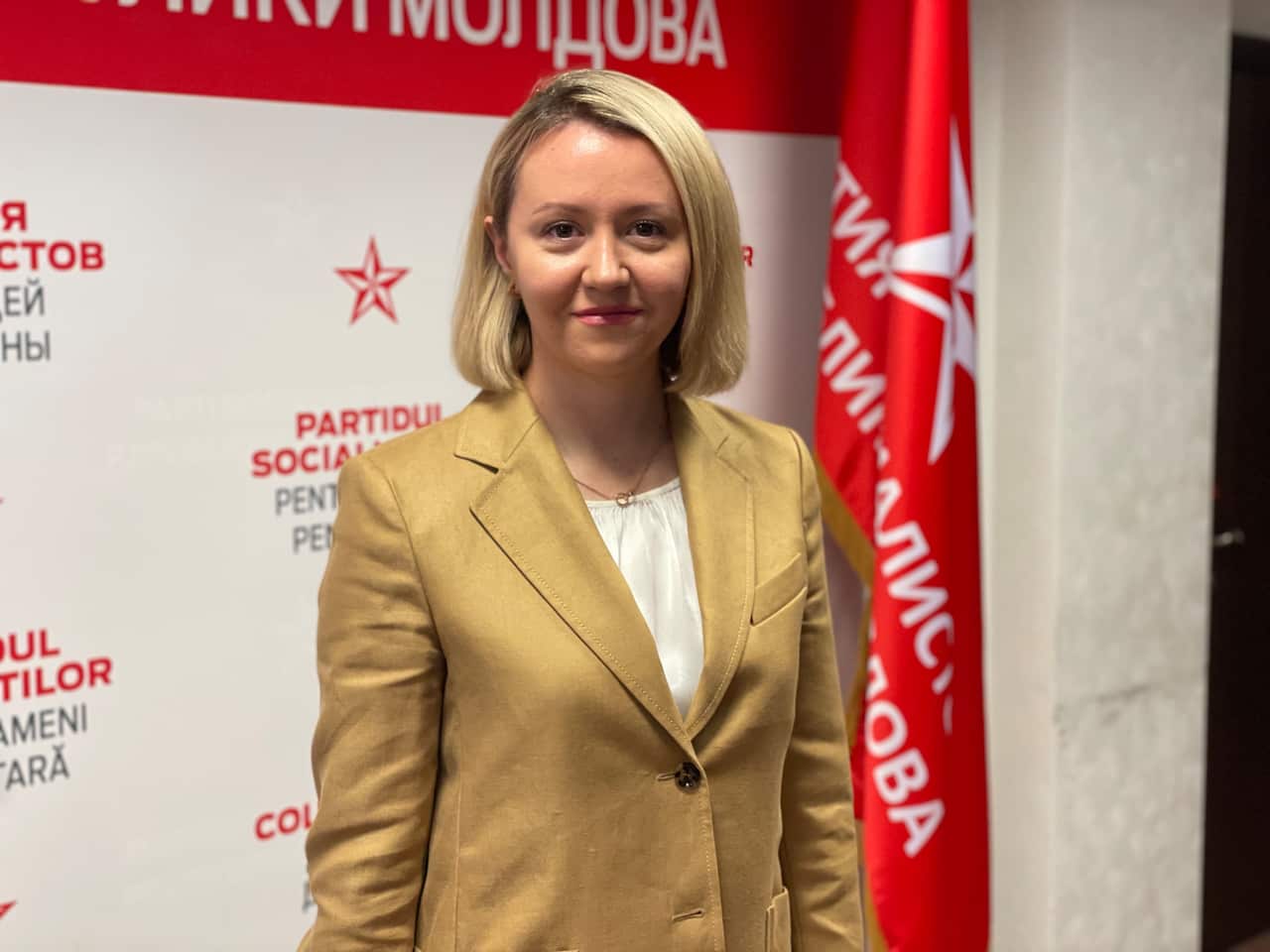 A young woman with blonde hair wearing a tan blazer standing in front of a red flag and poster.