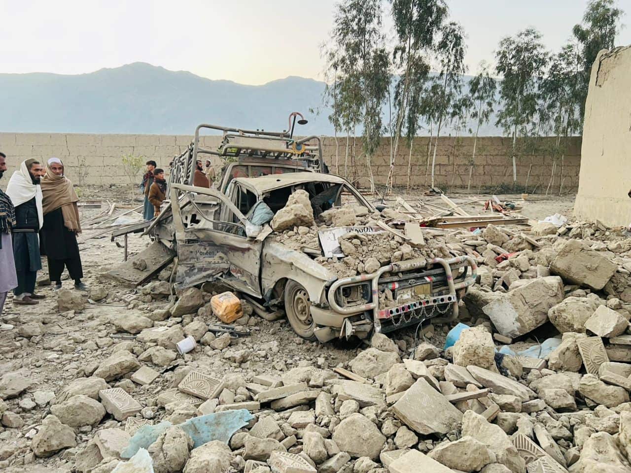 Rubble surrounding a burnt out car with people standing nearby