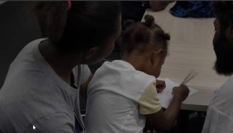 A photo taken from behind depicting a man and woman holding a small child, colouring a sheet of paper.