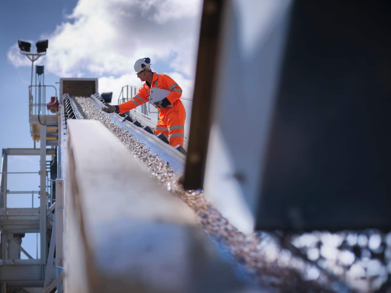 A man wearing a high-vis outfit and hard hat carries a clipboard as he lifts up rocks with a gloved hand as it moves down an outdoor conveyor belt