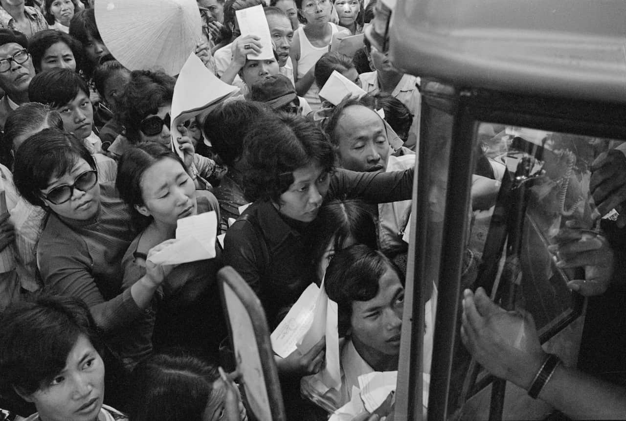 Crowds of people, holding papers, attempt to board a bus. 