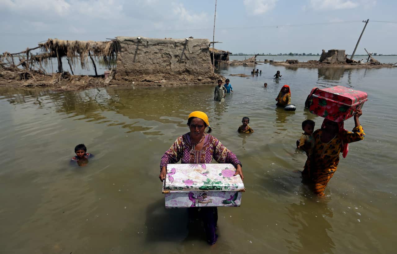 Women and children in waist-deep water carry their belongings in suitcases in a flooded landscape in Pakistan, with destroyed buildings in the background.
