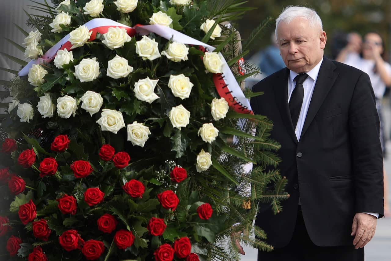A man with white hair and wearing a suit stands next to a big wreath of white and red roses