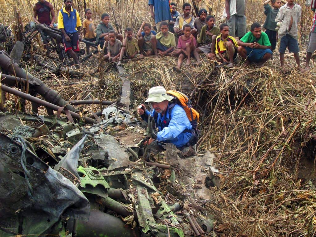 A man looks closely at the wreckage of a plane. People sit behind him watching.