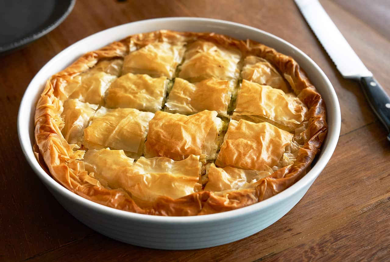 A golden filo pie scored into square pieces sits in a deep, round white pie dish.