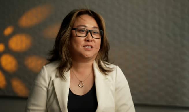 Uma mulher de óculos, vestindo uma jaqueta creme e uma blusa escura.