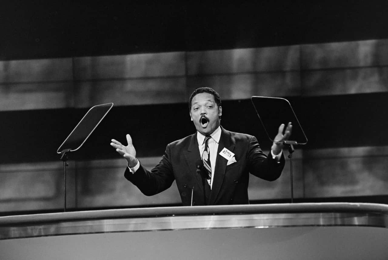 A black and white photograph of a black man in a suit speaking with his arms outstretched at a podium 