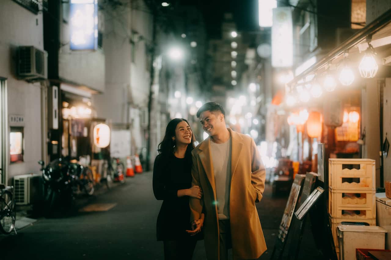 A young couple walks hand-in-hand down a narrow, glowing Tokyo alleyway at night, smiling warmly at each other amidst the blurred lights of storefronts and lanterns.