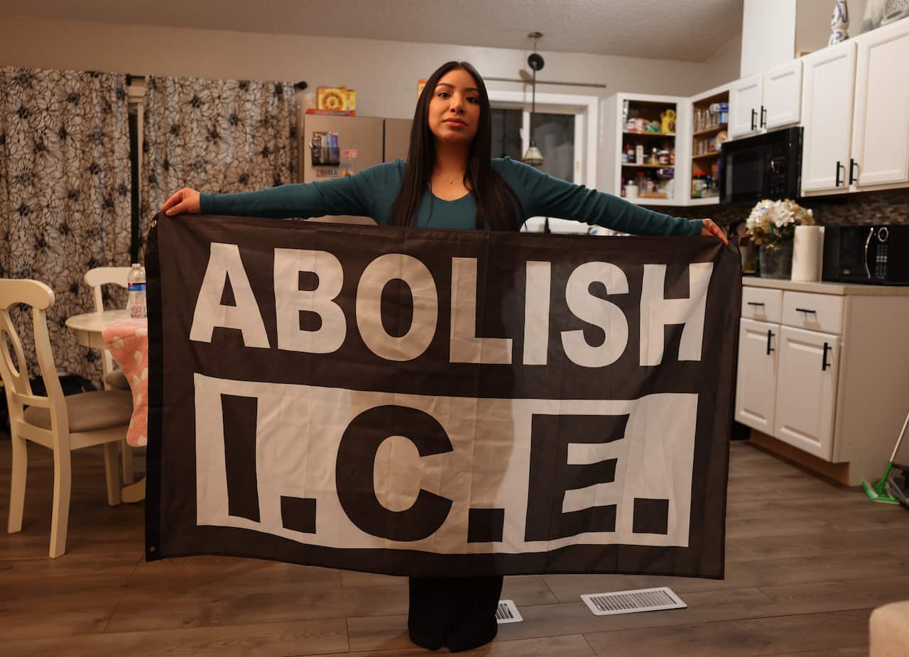 A woman stands in her kitchen holding a large banner which reads 'Abolish I.C.E' 