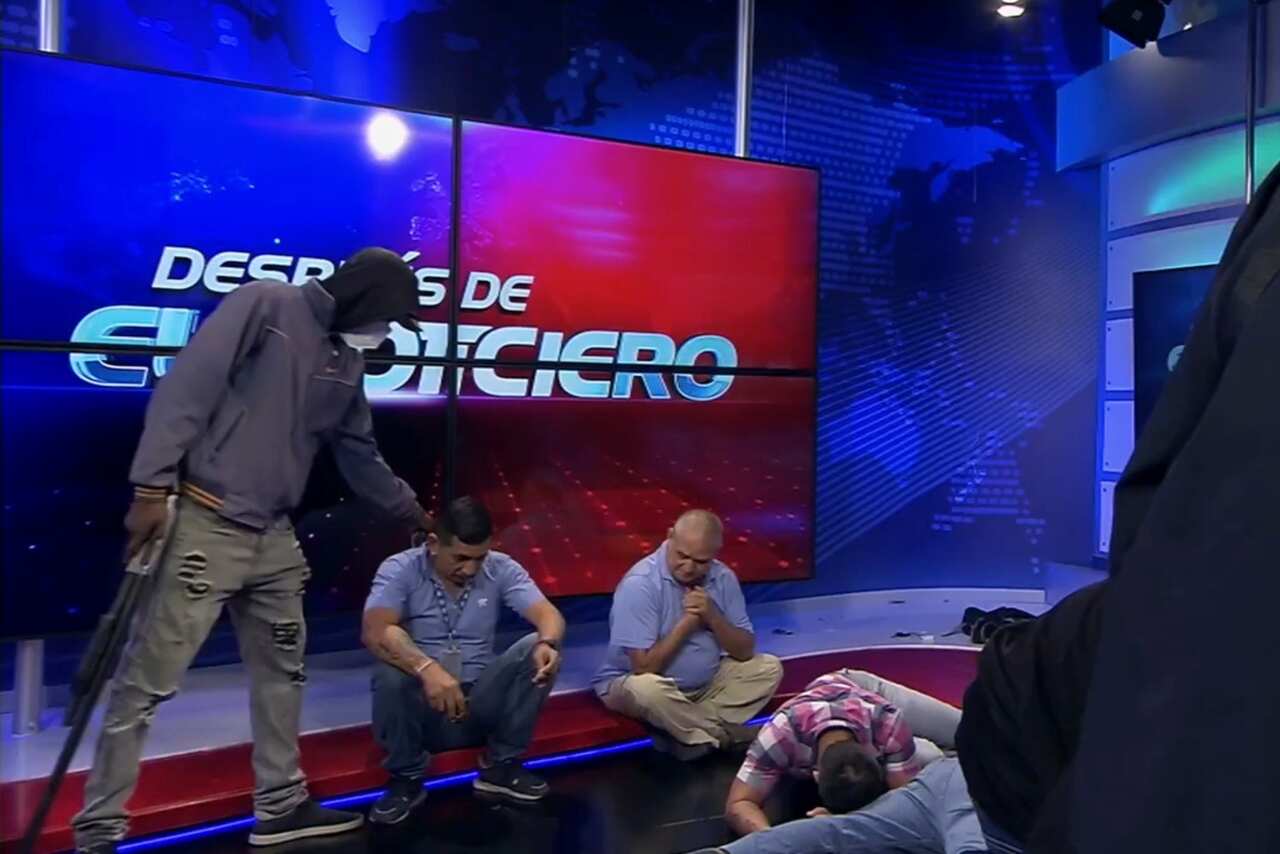 A person holding a gun and wearing a mask standing over journalists during a TV broadcast