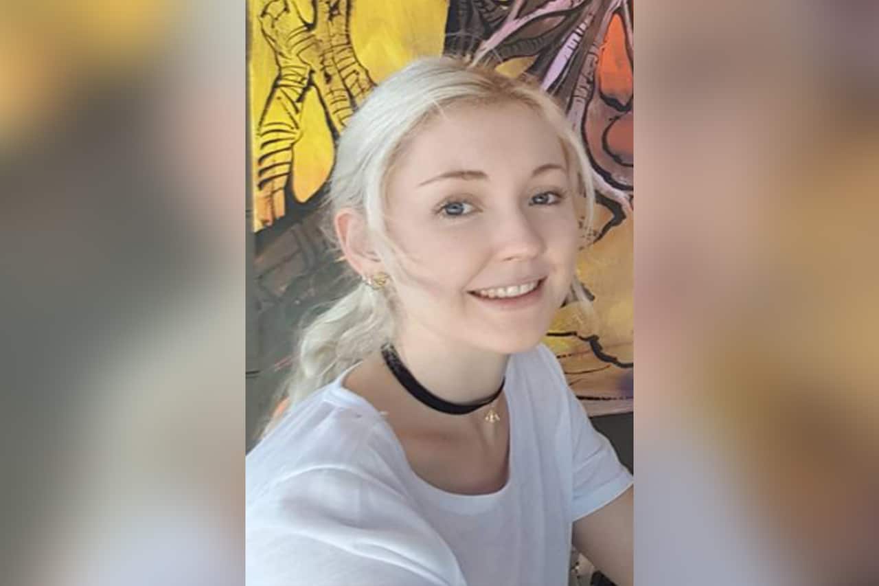 A young blonde woman wearing a white top and a black choker with her hair tied back.