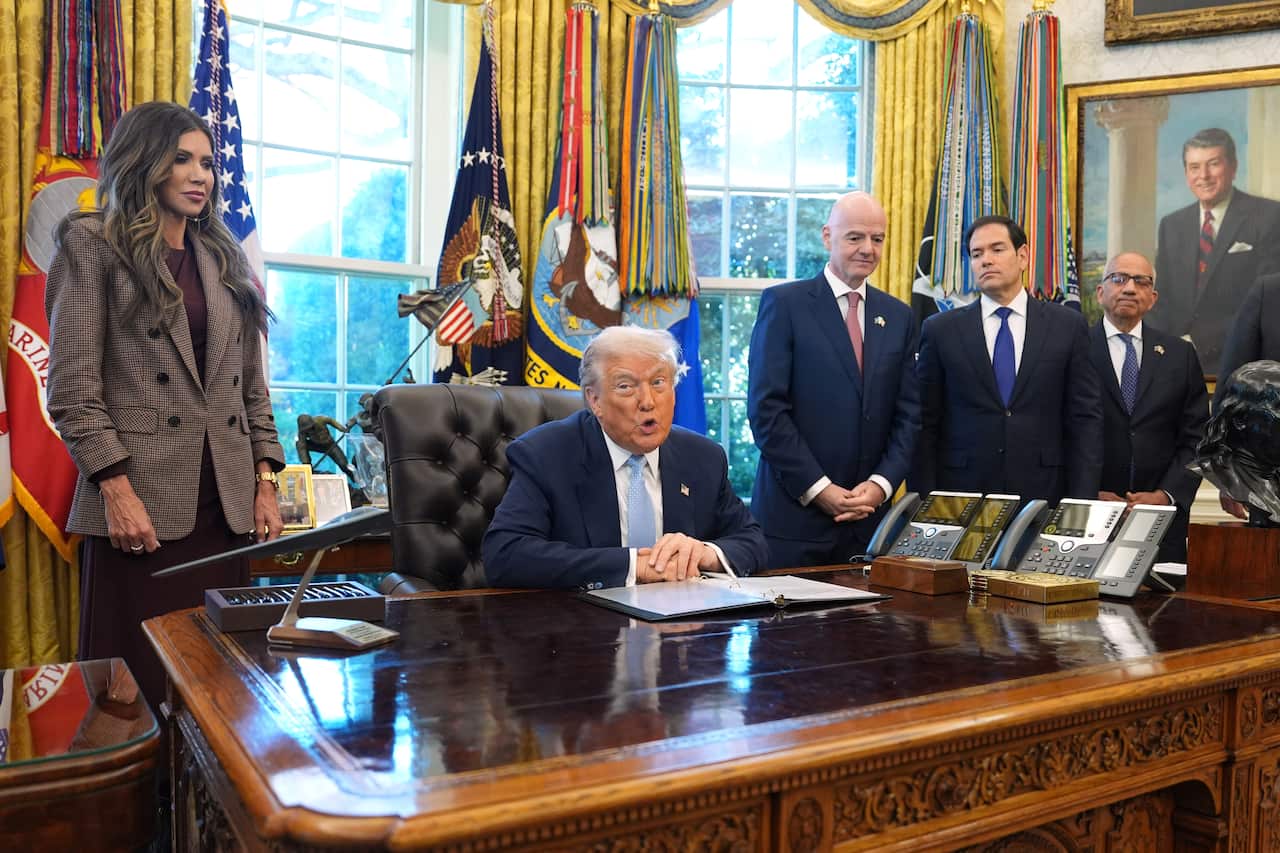 Donald Trump sitting in his chair in the Oval Office, flanked by a woman in a brown blazer, and three men in navy blazers.