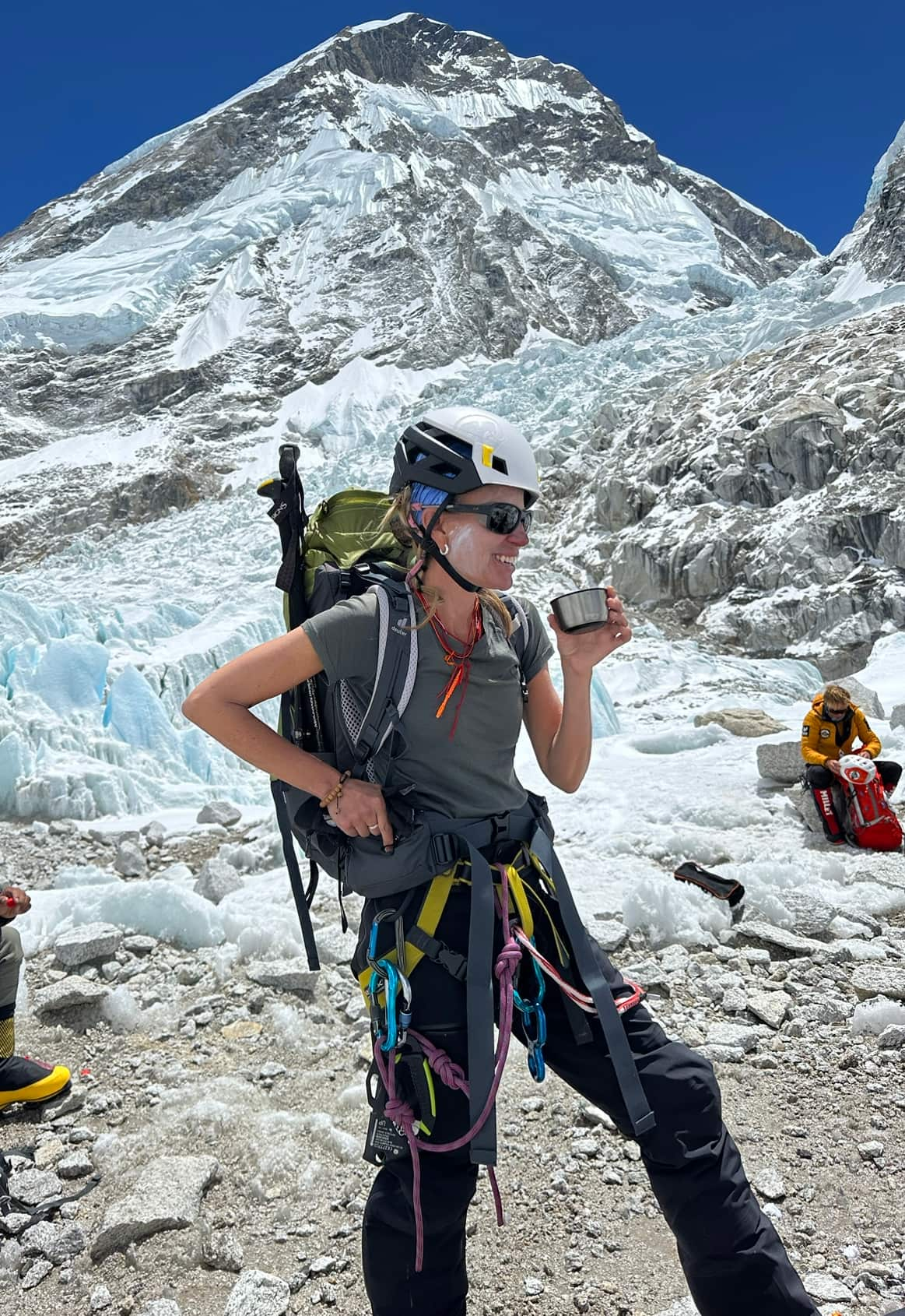 A woman wearing a helmet and mountaineering gear stands infront of a mountain.