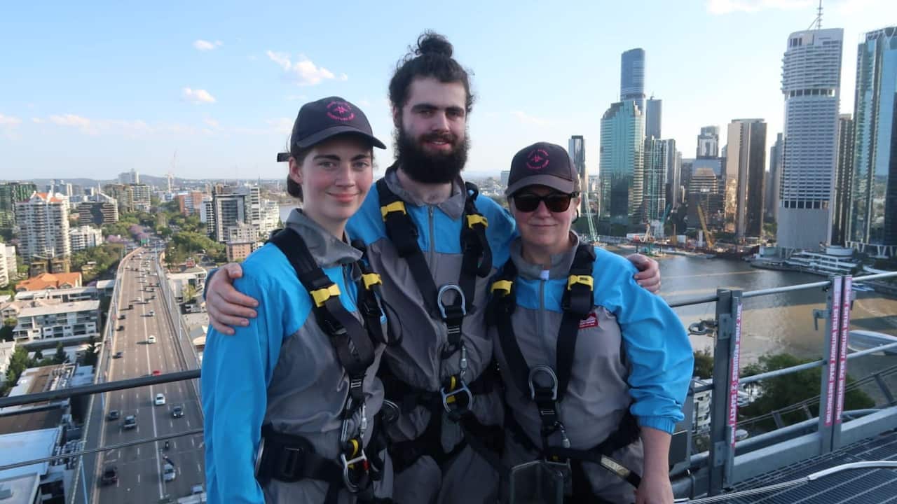 A teenage girl, a young man and a middle-aged woman in blue and grey jumpsuits with safety harnesses stand on a bridge with a city and river in the background.