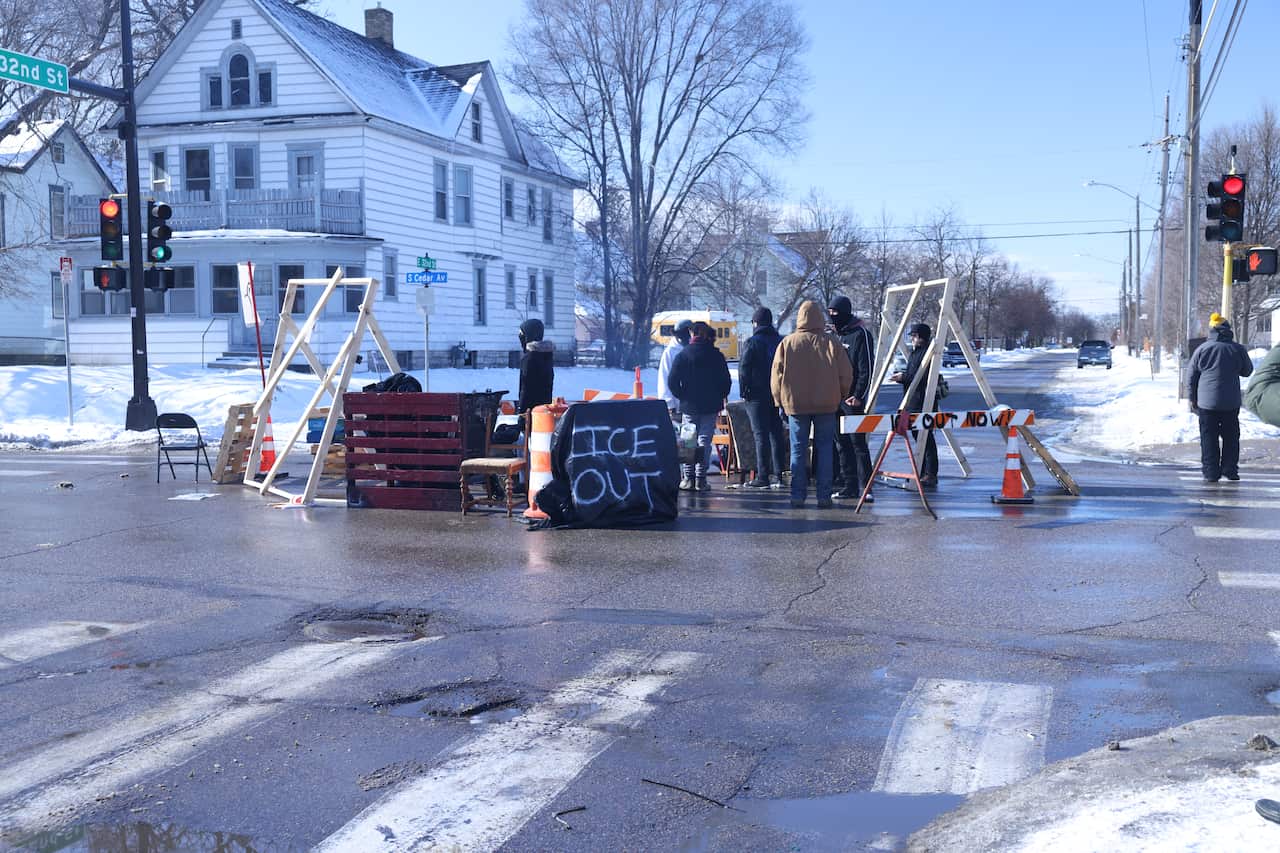 A group of people stand in the middle of a street intersection. They have created a barricade using cones, chairs and wooden structures. They have a sign reading ICE OUT.