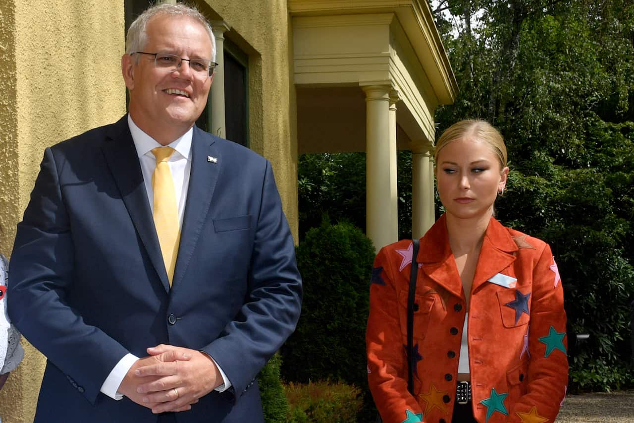 Scott Morrison and Grace Tame, standing next to each other. Morrison is wearing a blue suit, Tame is wearing a red blazer with coloured stars. She has a tense expression on her face.