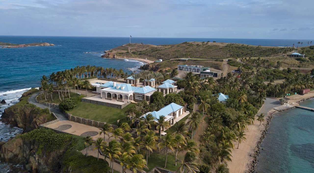 Aerial view of a secluded private island with a large waterfront estate, palm trees, multiple villas with blue roofs, and surrounding turquoise waters.
