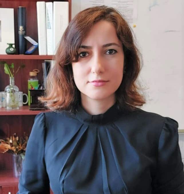 Una mujer con cabello castaño hasta los hombros y una camisa azul está sentada frente a una estantería.