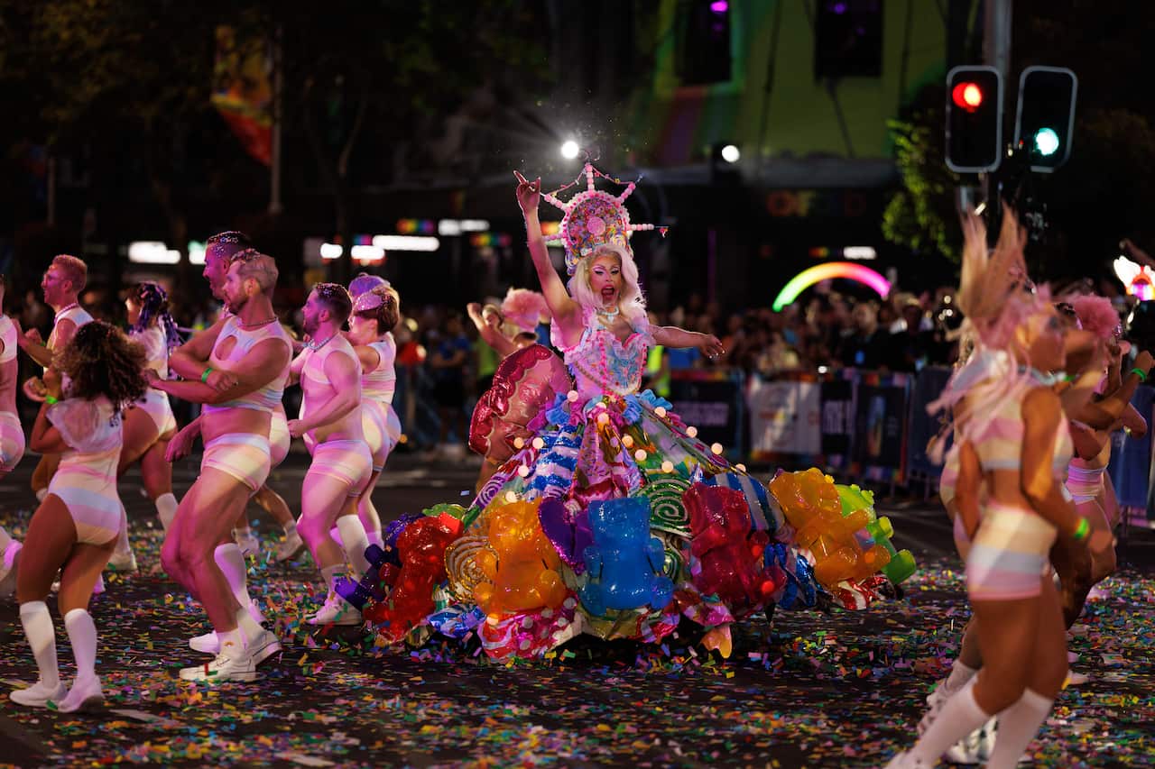 Una drag queen actúa en medio de una calle cubierta de confeti. Su enorme y colorida falda está decorada con enormes flores translúcidas. "ositos de goma" y decoraciones con temas de dulces. Los rodean bailarines con trajes de colores pastel.
