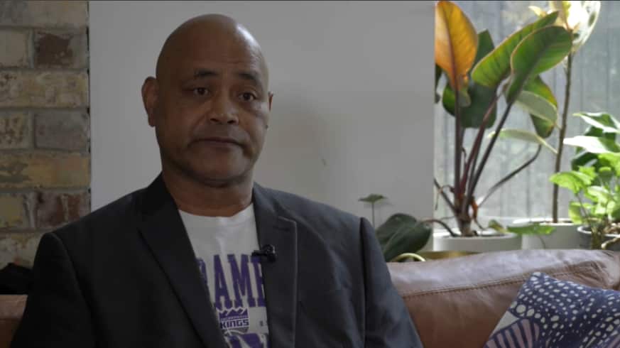 A middle-aged Fijian Australian man wearing a white shirt and black blazer sits on a lounge.
