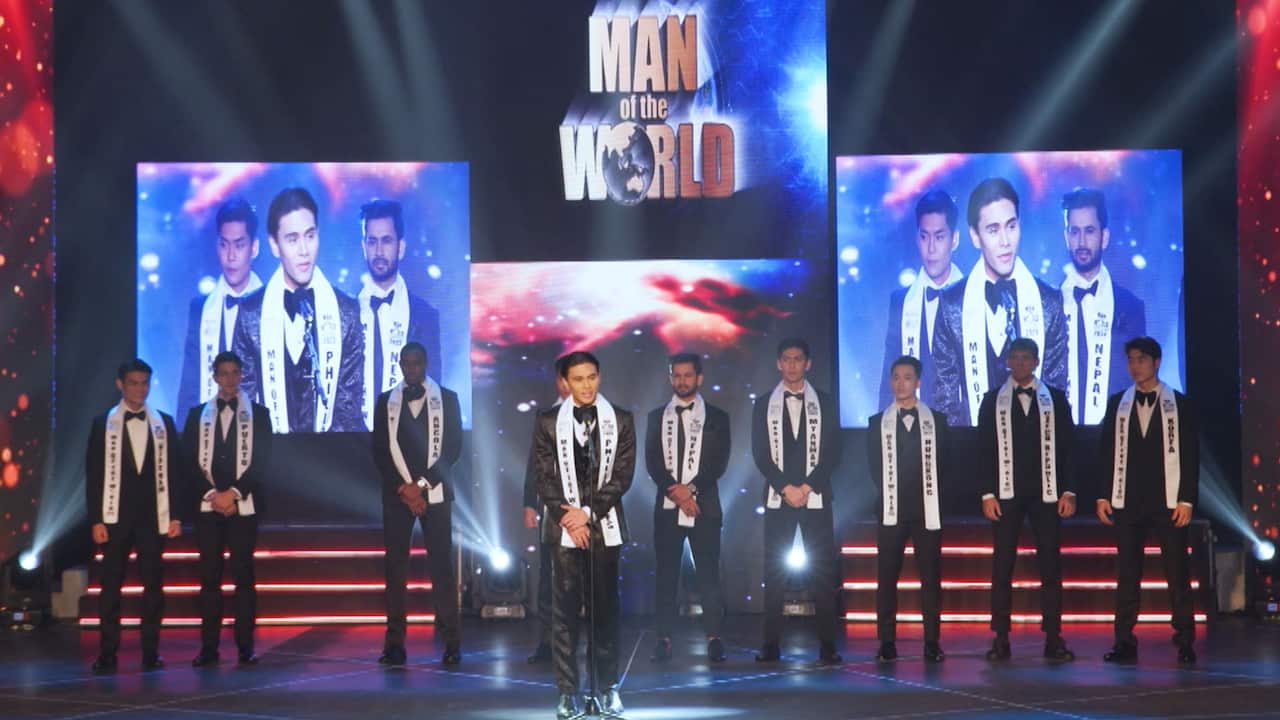 Ten men in tuxedos and white pageant sashes standing on stage while one of them speaking into the microphone during a male beauty competition