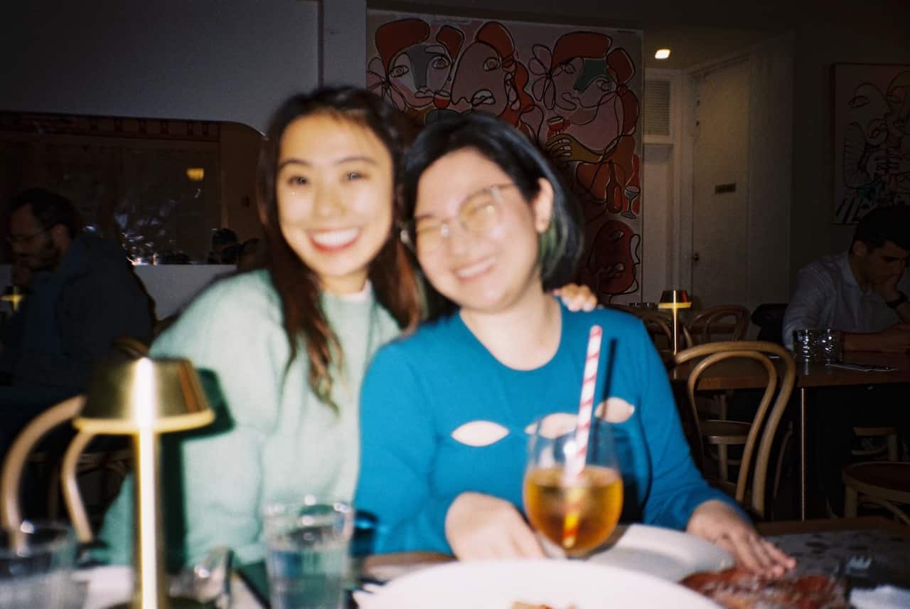 Two women seated next to each other at a restaurant smile, one with their arm on the other's shoulder.