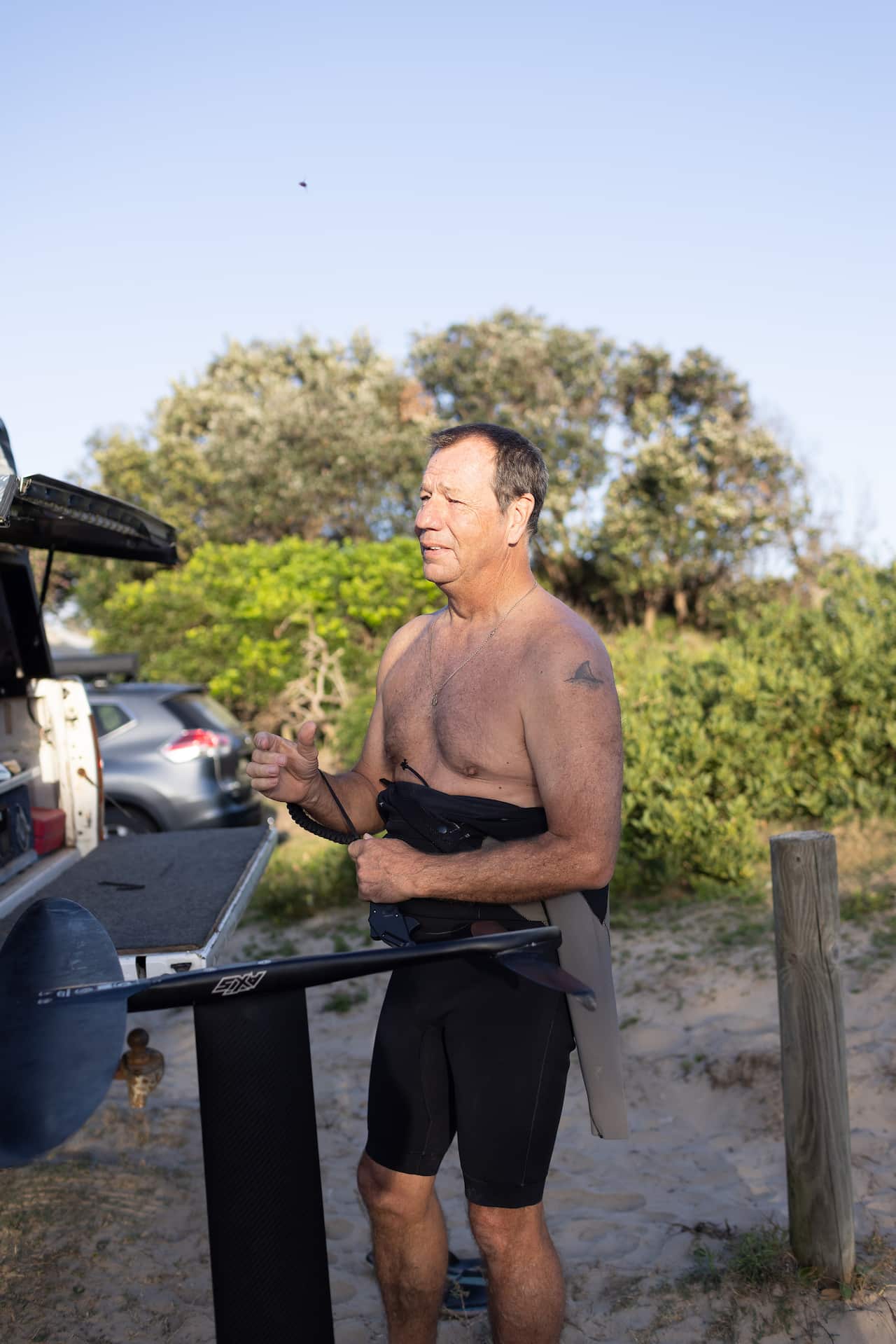 A man with a wetsuit on the lower half of his body stands near several parked cars. 