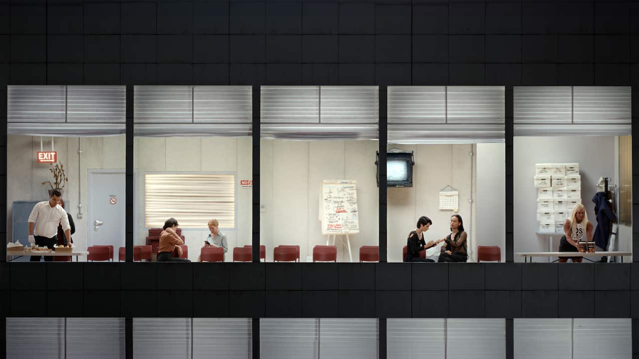 The view through the wall of office windows shows a series of seated and standing people.  The lighting suggests it might be dark.