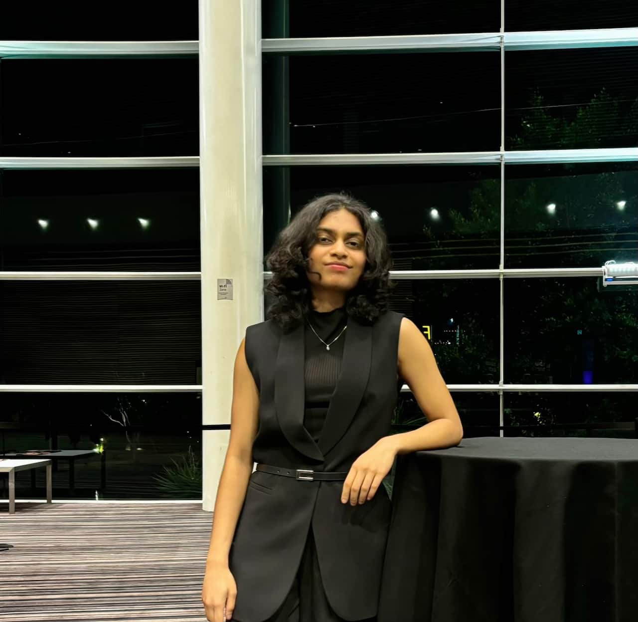 A woman in a black dress leans against a table and smiles at the camera.