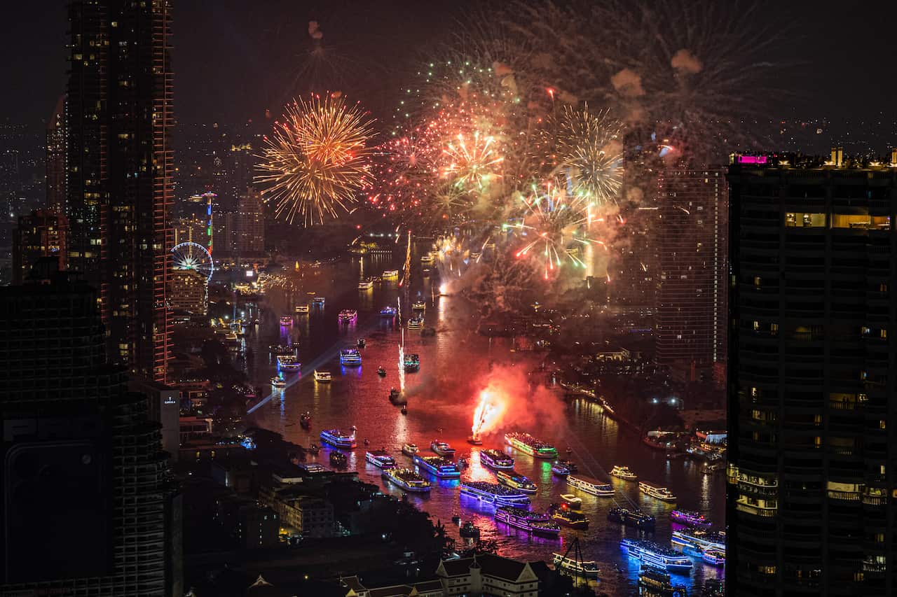 Fireworks over a river winding through a city. Boats are lit up with colourful lights on the water.