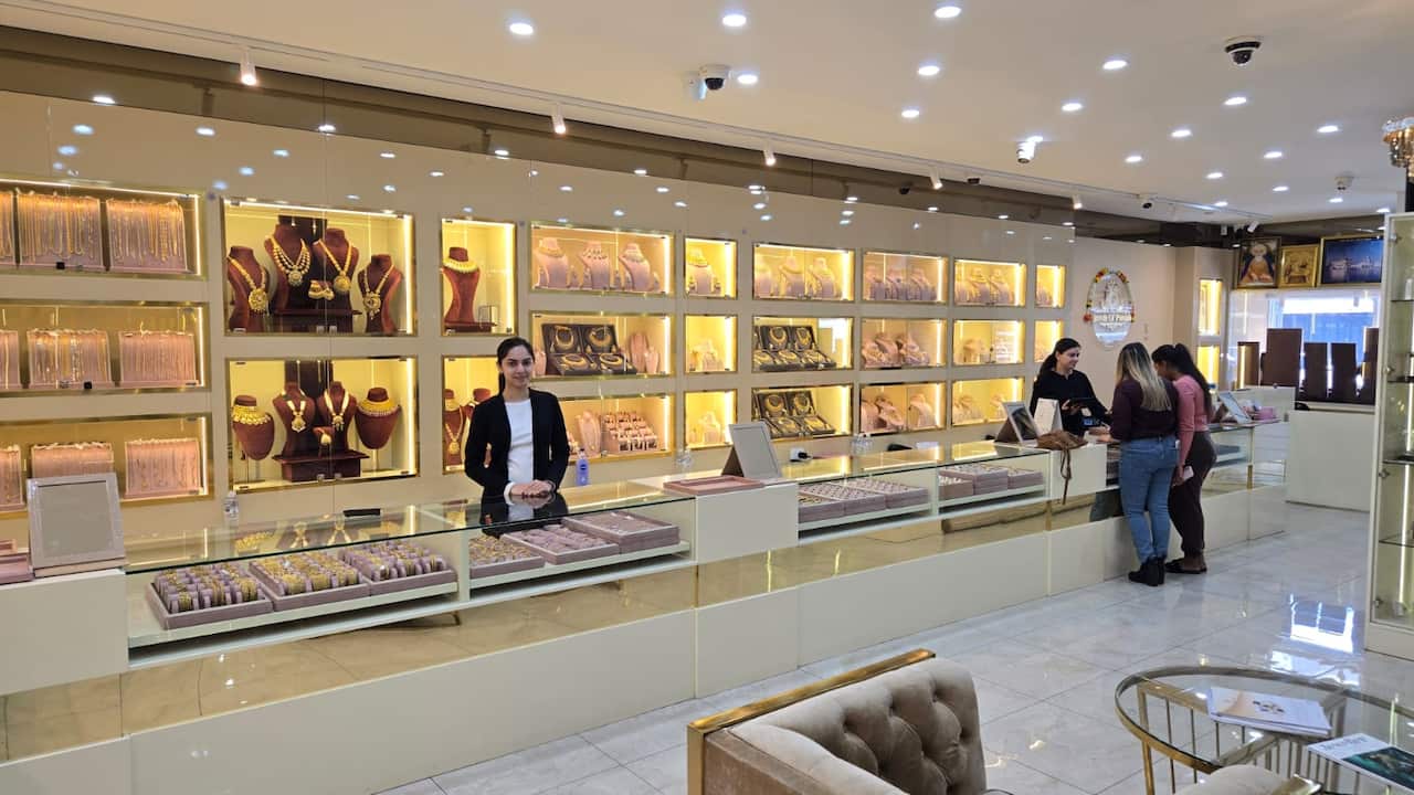 A woman standing behind a glass counter filled with gold jewellery inside a brightly lit jewellery showroom. There's another saleswoman attending to two customers standing at a distance.