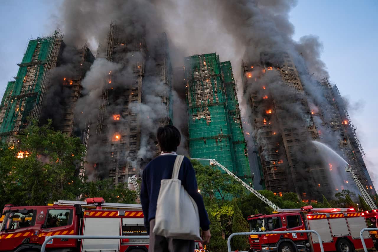 A person stands with their back to the camera, watching thick smoke and flames engulf several high-rise buildings under construction as firefighters battle the massive blaze with water cannons.