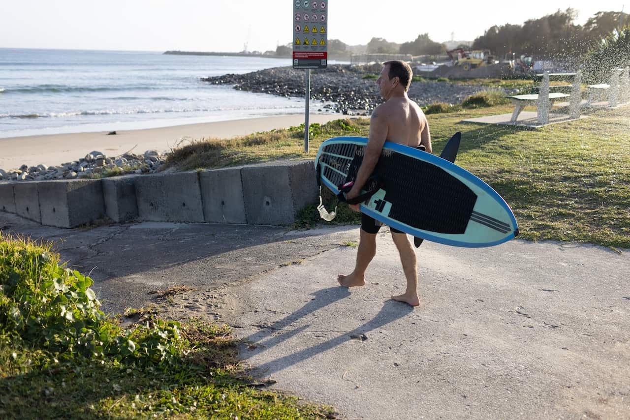 A man in a wetsuit carries a surfboard while walking on a concrete path towards a beach. 