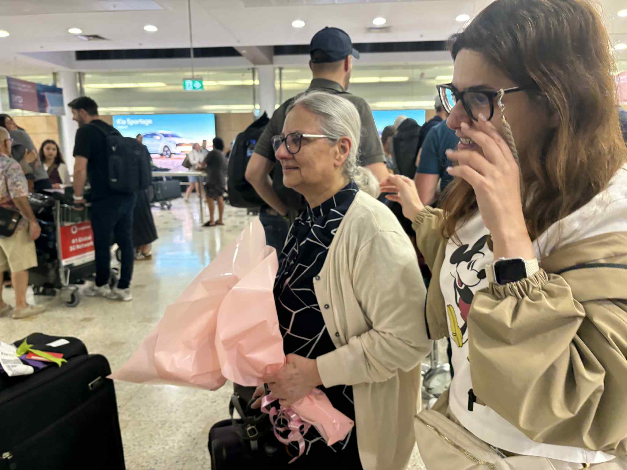 An older woman with white hair tied back in a low ponytail, holding a bunch of flowers wrapped in light pink cellophane, walking through an airport with many people in the background. Another woman is walking beside her. 