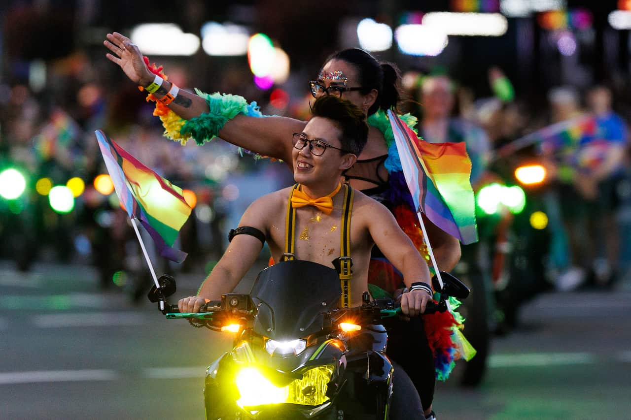 Una persona feliz con gafas y pajarita amarilla monta una motocicleta en un desfile nocturno. Hay dos banderas de Progress Pride en el manillar. Detrás de ellos, un pasajero con brillo en el rostro saluda a la multitud, envuelto en una boa de plumas voluminosa y colorida.