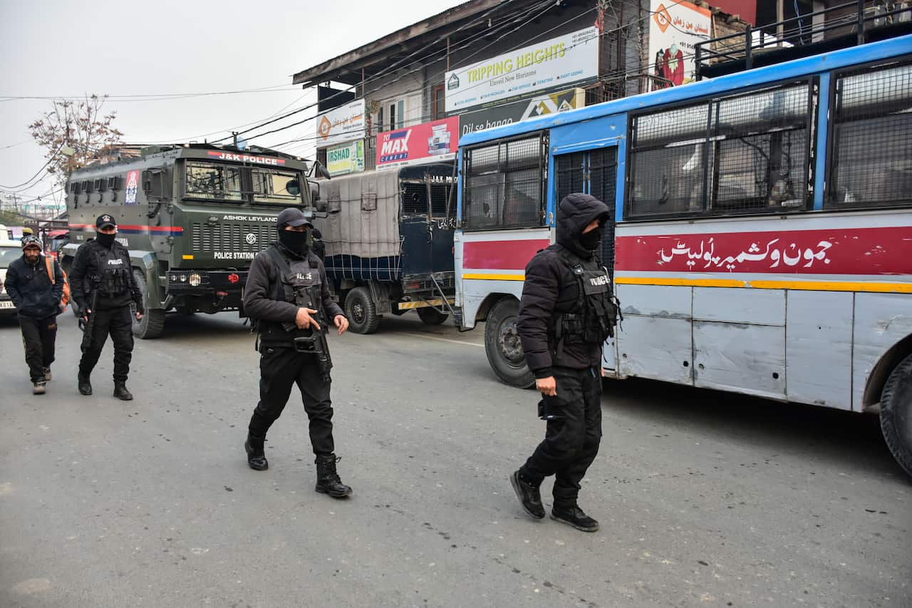 Several masked police officers in black tactical gear patrol a street alongside an armoured police truck and a caged bus, with other pedestrians and vehicles in the background.