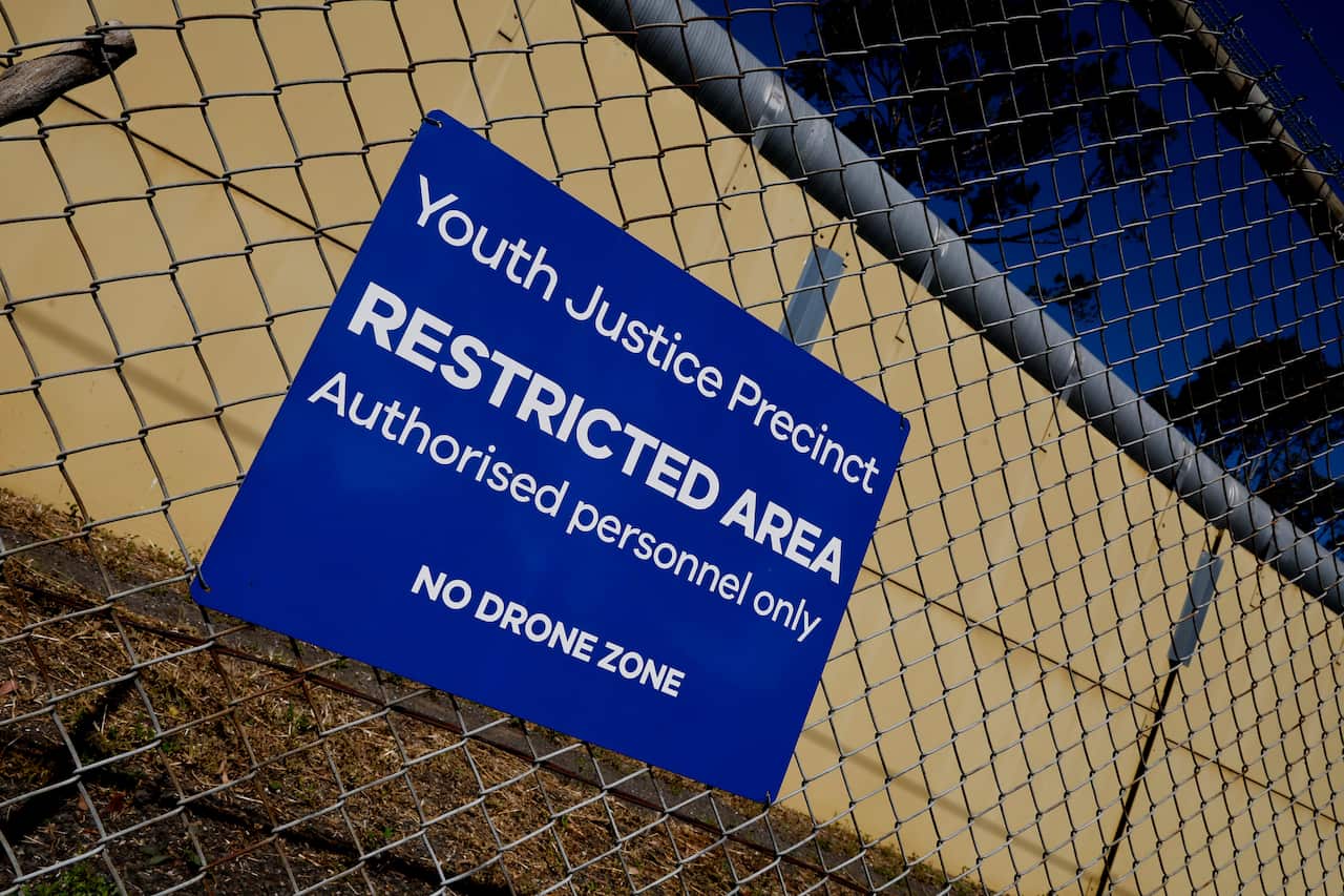 A blue 'Youth Justice Precinct' sign on a wire fence around a yellow building.