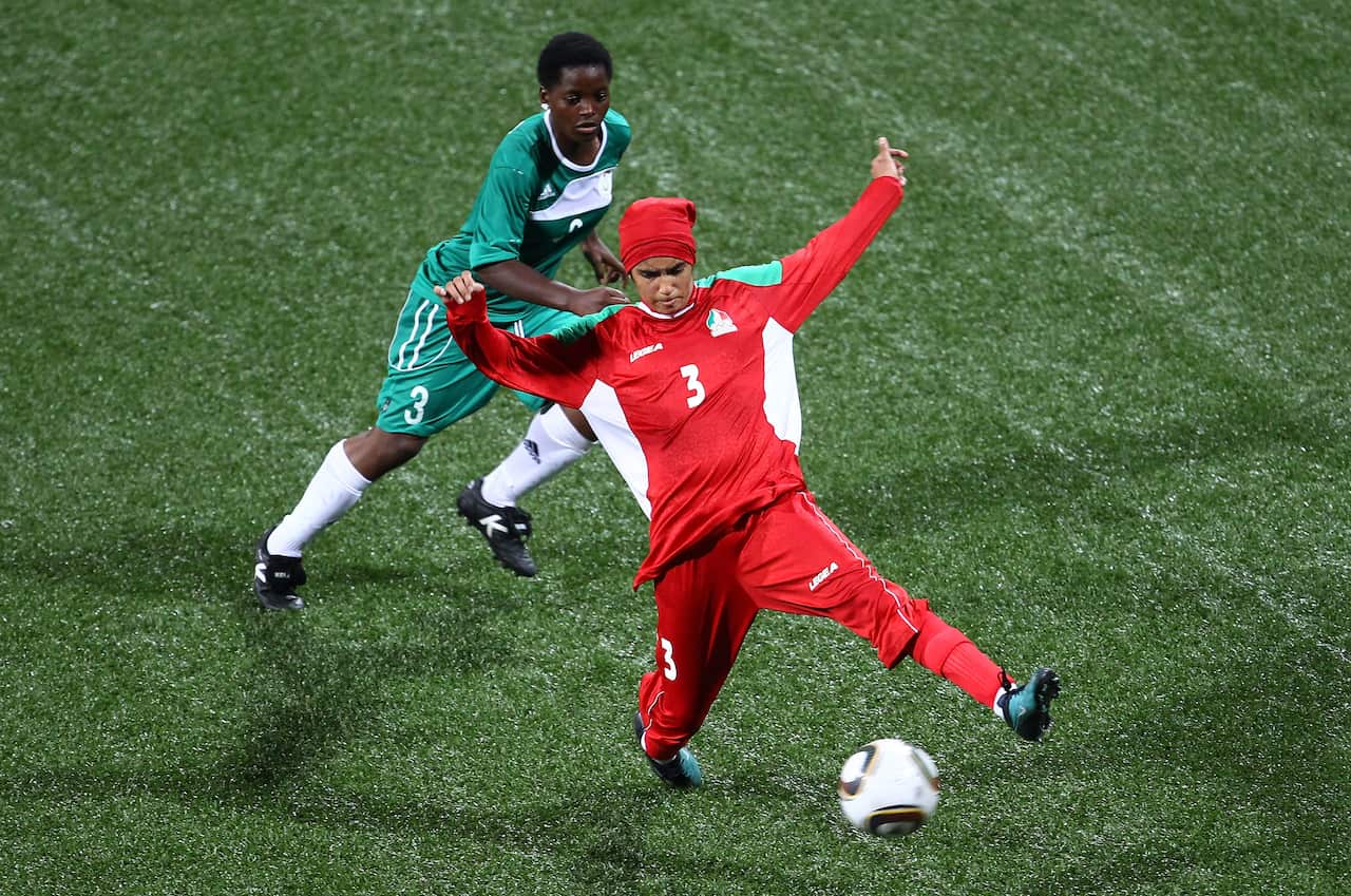 A woman in red uniform kicks a soccer ball, while a woman in green behind her tries to defend