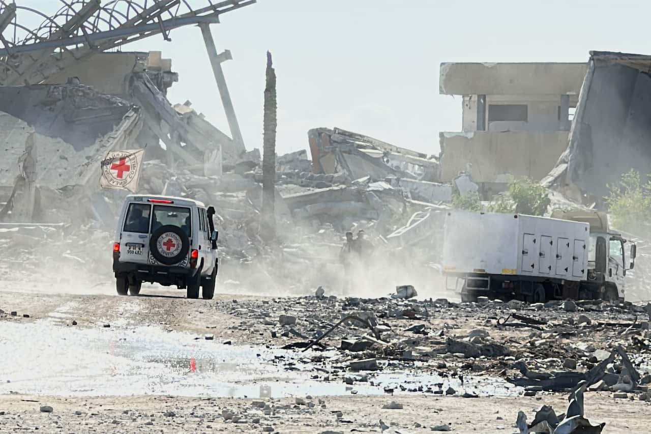 A Red Cross vehicle passes through a dirt road flanked by destroyed buildings and rubble. There's a white truck ahead.
