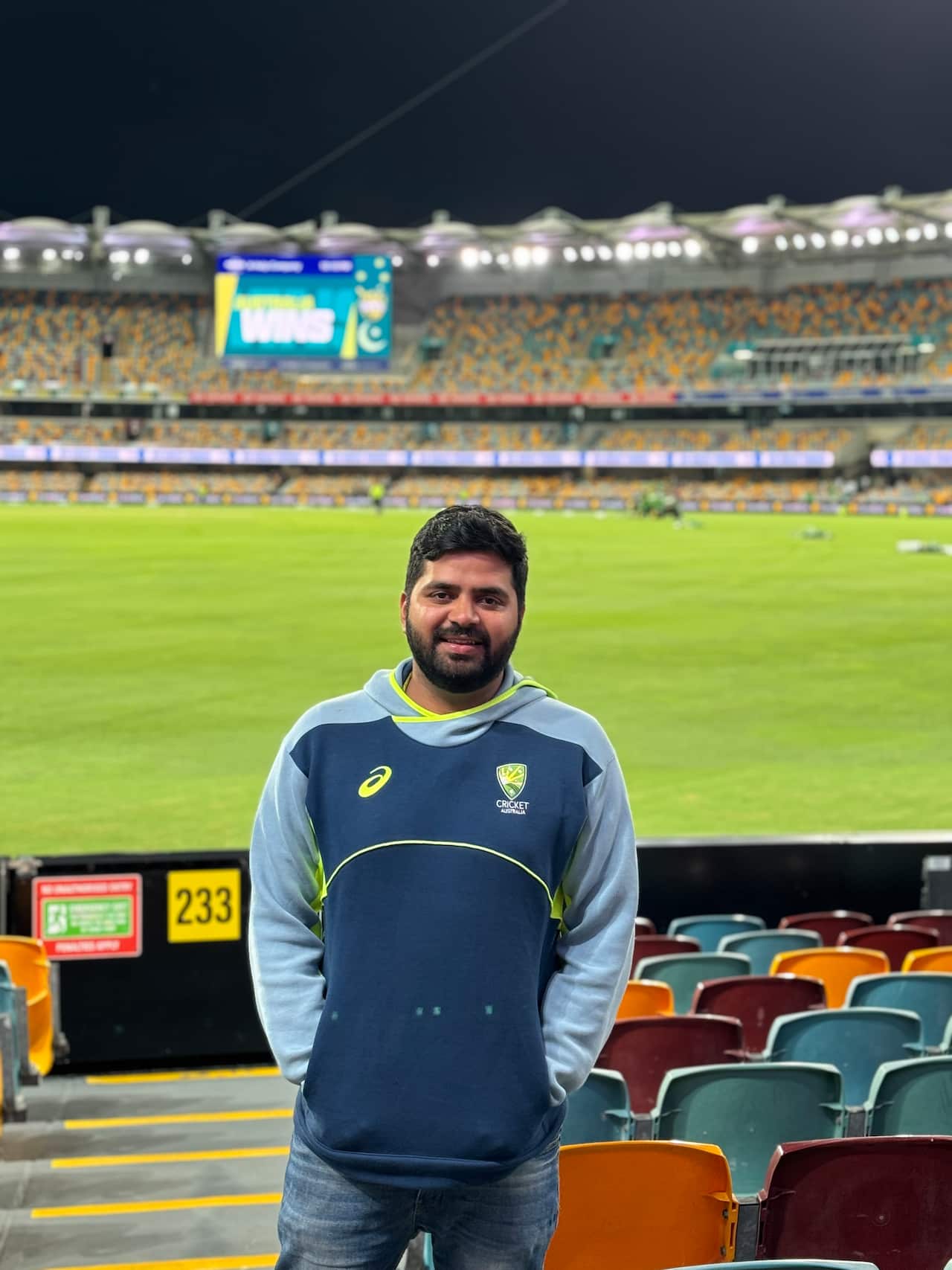A man in a navy and grey hoodie stands in front of colourful stadium seating and a brightly lit field of a large cricket arena at night.